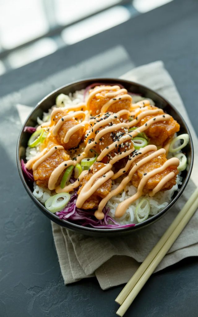 Crispy golden salmon bites piled over white jasmine rice in a dark ceramic bowl drizzled with sriracha mayo and topped with sesame seeds and green onions