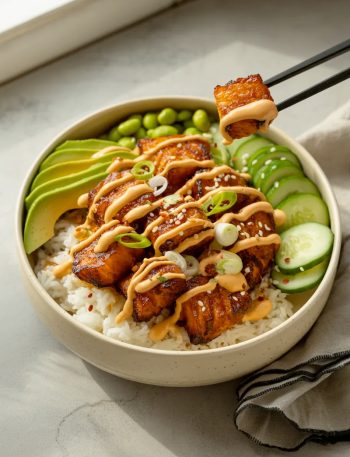Crispy sriracha glazed salmon bites over white rice in a speckled ceramic bowl with sliced avocado edamame cucumber sesame seeds and sriracha mayo drizzle with black chopsticks lifting one piece