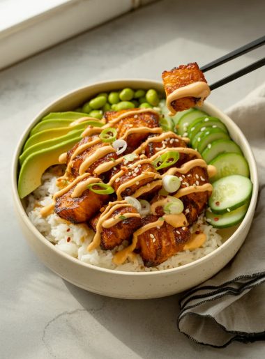 Crispy sriracha glazed salmon bites over white rice in a speckled ceramic bowl with sliced avocado edamame cucumber sesame seeds and sriracha mayo drizzle with black chopsticks lifting one piece