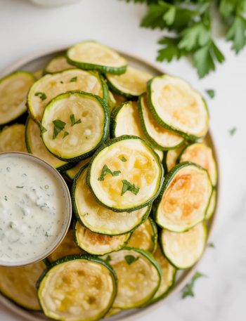 A white plate piled with golden crispy baked zucchini chips garnished with fresh parsley and served with a creamy dipping sauce