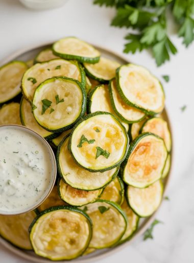 A white plate piled with golden crispy baked zucchini chips garnished with fresh parsley and served with a creamy dipping sauce