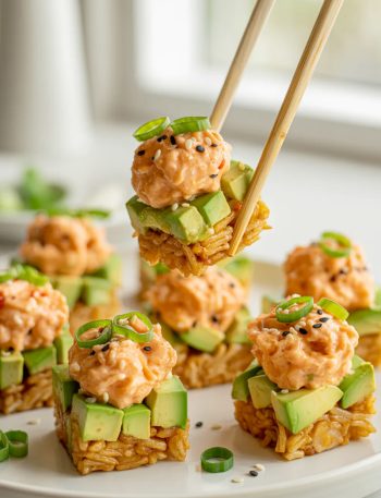 Several crispy golden rice bites topped with creamy spicy salmon mixture diced avocado black sesame seeds and sliced green onions arranged on a white plate with chopsticks picking up one piece