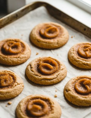 Six golden brown cinnamon brown butter cookies arranged on white parchment paper showing a beautiful caramelized cinnamon swirl pattern on top of each cookie