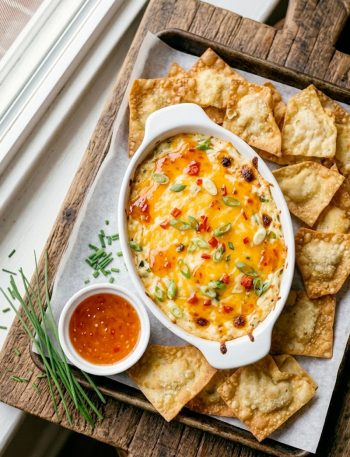 Overhead view of baked crab rangoon dip in a white oval dish topped with sweet chili sauce and green onions surrounded by crispy wonton chips