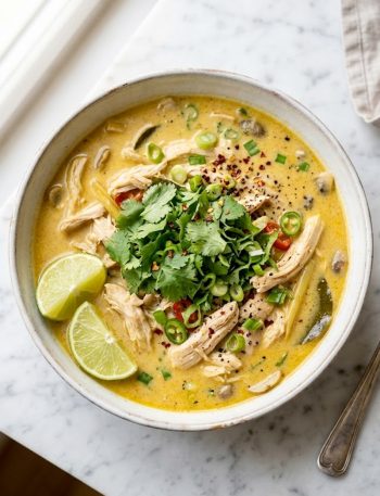Overhead shot of a bowl of creamy Thai coconut chicken soup garnished with fresh cilantro and lime wedges on a white surface
