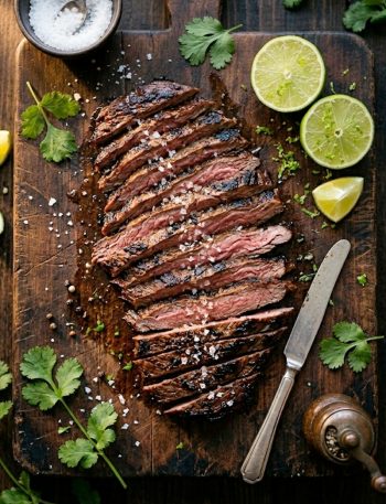 Thinly sliced marinated skirt steak with a deep charred crust fanned out on a wooden cutting board with fresh cilantro and lime wedges