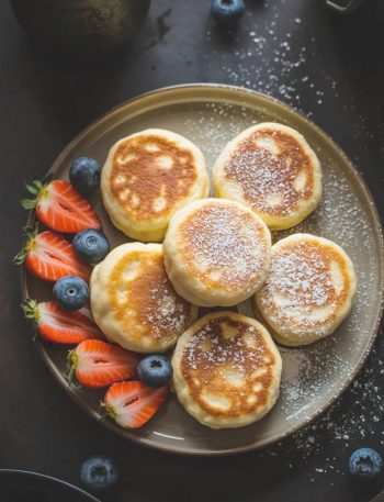 Overhead view of a stack of small golden fluffy cottage cheese pancakes dusted with powdered sugar served alongside fresh sliced strawberries and blueberries on a dark ceramic plate