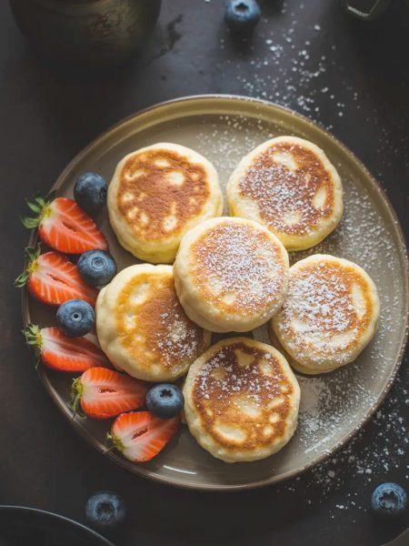 Overhead view of a stack of small golden fluffy cottage cheese pancakes dusted with powdered sugar served alongside fresh sliced strawberries and blueberries on a dark ceramic plate