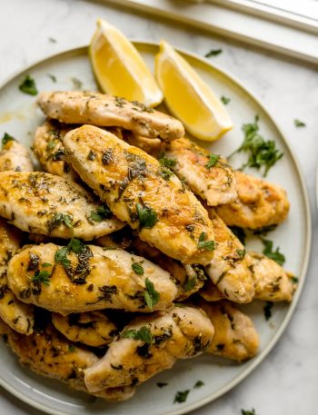 A white plate piled high with golden juicy Greek chicken tenders garnished with fresh chopped parsley and served with lemon wedges on a marble surface