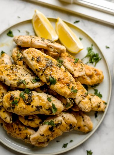 A white plate piled high with golden juicy Greek chicken tenders garnished with fresh chopped parsley and served with lemon wedges on a marble surface