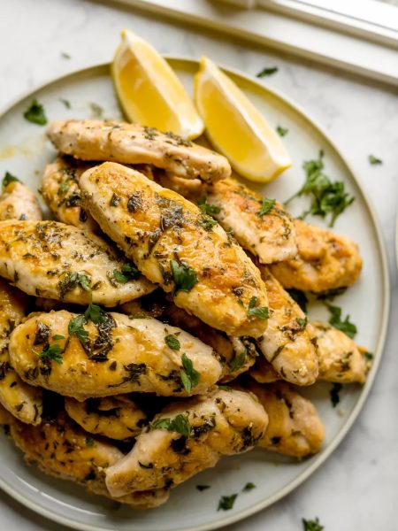 A white plate piled high with golden juicy Greek chicken tenders garnished with fresh chopped parsley and served with lemon wedges on a marble surface