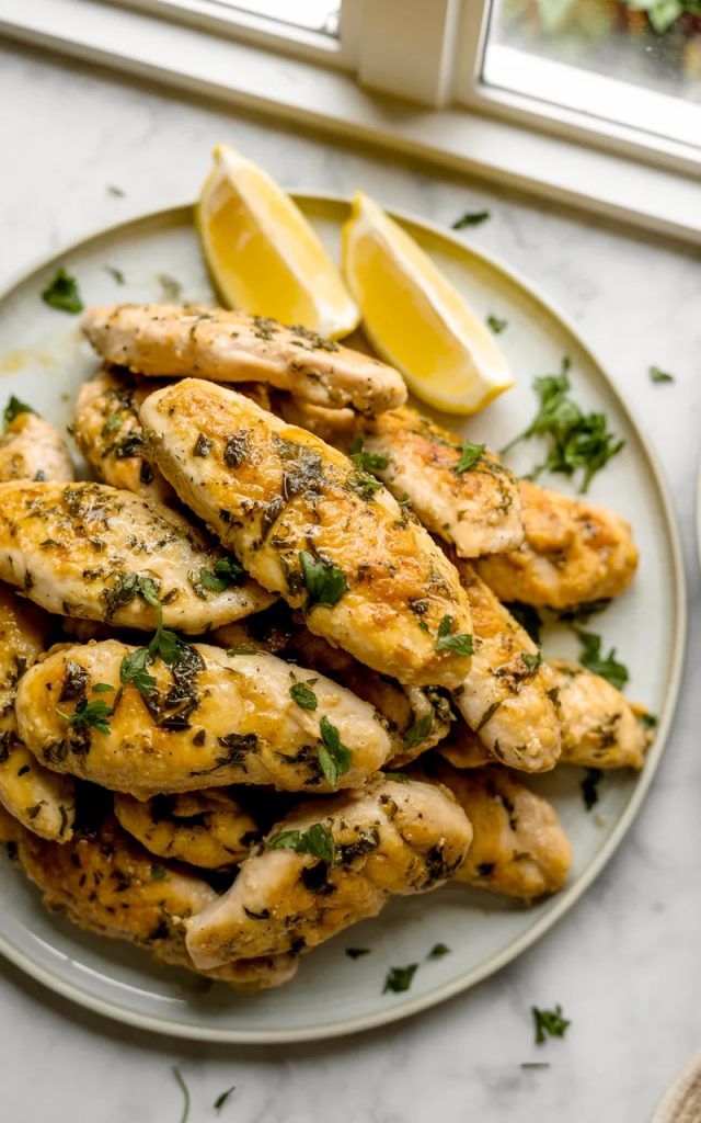 A white plate piled high with golden juicy Greek chicken tenders garnished with fresh chopped parsley and served with lemon wedges on a marble surface