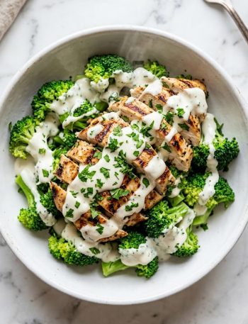 A grilled chicken and broccoli bowl drizzled with creamy garlic sauce and garnished with fresh parsley in a white bowl on a marble surface