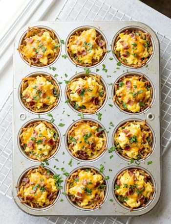 A close up overhead shot of golden crispy hash brown egg cups filled with fluffy scrambled eggs, crumbled bacon, and melted cheddar cheese garnished with fresh parsley in a muffin tin