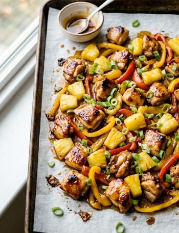 Hawaiian chicken sheet pan with caramelized pineapple chunks, red and yellow bell peppers, and green onions on a dark roasting pan