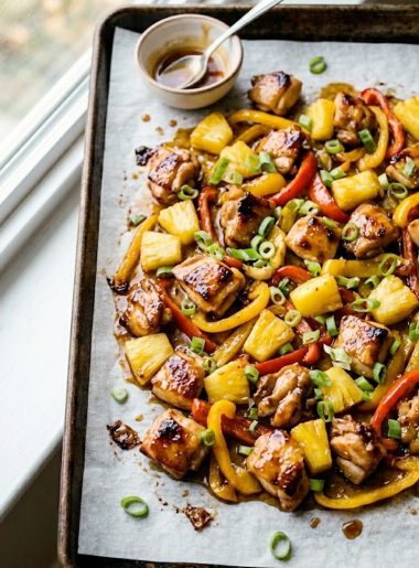 Hawaiian chicken sheet pan with caramelized pineapple chunks, red and yellow bell peppers, and green onions on a dark roasting pan