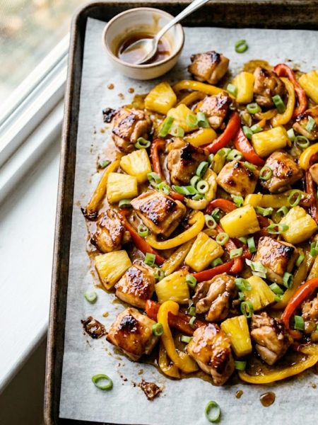 Hawaiian chicken sheet pan with caramelized pineapple chunks, red and yellow bell peppers, and green onions on a dark roasting pan