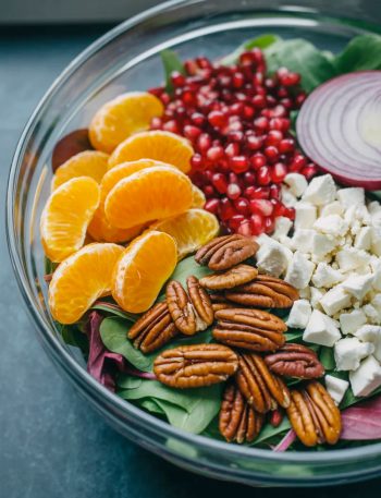 A large clear glass bowl filled with a stunning healthy winter salad showing bright orange mandarin segments ruby red pomegranate seeds golden candied pecans white crumbled feta and dark green mixed greens before tossing