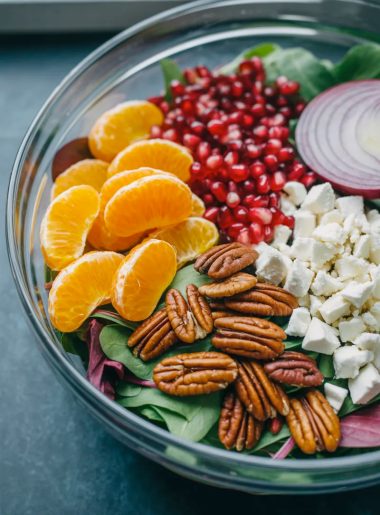 A large clear glass bowl filled with a stunning healthy winter salad showing bright orange mandarin segments ruby red pomegranate seeds golden candied pecans white crumbled feta and dark green mixed greens before tossing