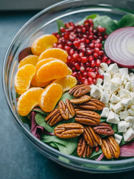 A large clear glass bowl filled with a stunning healthy winter salad showing bright orange mandarin segments ruby red pomegranate seeds golden candied pecans white crumbled feta and dark green mixed greens before tossing