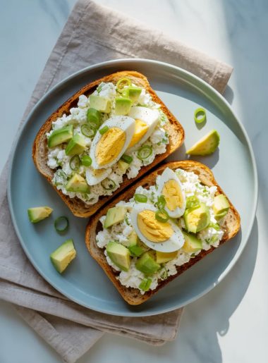 Two slices of toasted whole grain bread topped with creamy cottage cheese egg salad, diced avocado, and sliced green onions on a light gray ceramic plate