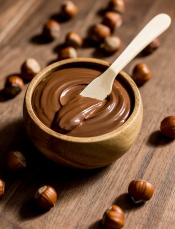 A wooden bowl filled with smooth homemade chocolate hazelnut spread with a butter knife resting inside surrounded by whole hazelnuts on a dark wooden surface