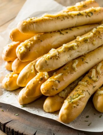 A pile of golden homemade Olive Garden style breadsticks on white parchment paper glistening with garlic butter and sprinkled with dried parsley and herbs