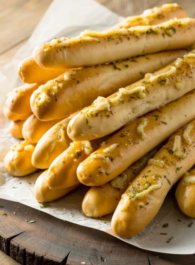 A pile of golden homemade Olive Garden style breadsticks on white parchment paper glistening with garlic butter and sprinkled with dried parsley and herbs