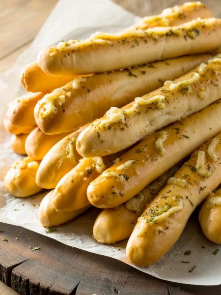 A pile of golden homemade Olive Garden style breadsticks on white parchment paper glistening with garlic butter and sprinkled with dried parsley and herbs