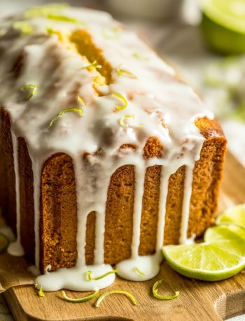 A whole key lime pound cake on a rustic wooden cutting board with thick white key lime glaze dripping down the sides and fresh lime zest scattered on top with one slice cut showing the dense moist golden interior