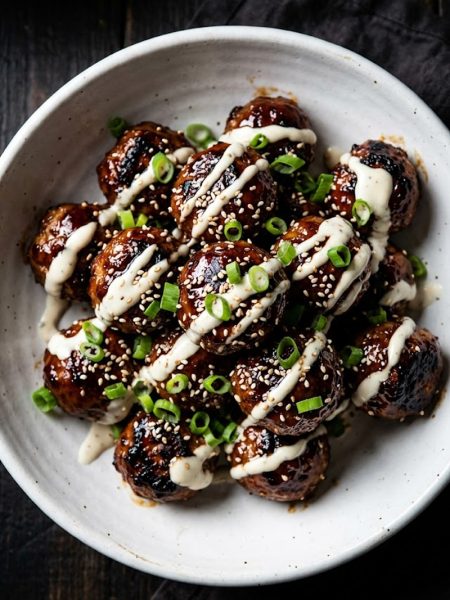 Close-up of glossy Korean BBQ meatballs in a white bowl with creamy gochujang drizzle and sliced green onions