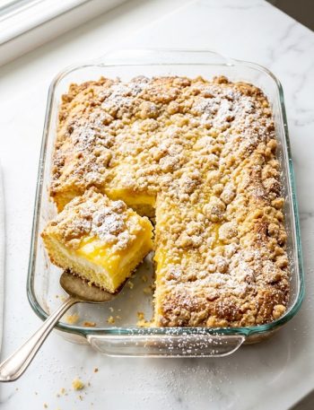A glass baking dish filled with golden baked lemon cream cheese dump cake with a crispy buttery topping, one square slice lifted to show the creamy lemon and cream cheese layers underneath