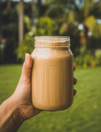 A hand holding a large mason jar filled with a thick creamy coffee banana smoothie against a blurred green outdoor background in natural morning light
