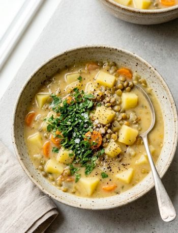 Overhead view of a rustic ceramic bowl filled with thick hearty lentil potato soup with chunky carrots, potato pieces, and green lentils, garnished with fresh parsley, styled on a light grey surface with a linen napkin