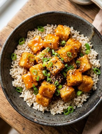 A bowl of crispy orange tofu served over brown rice, garnished with sliced green onions and sesame seeds