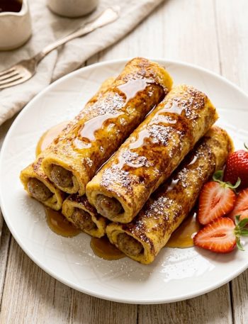 Golden brown sausage French toast roll-ups stacked on a white plate drizzled with maple syrup