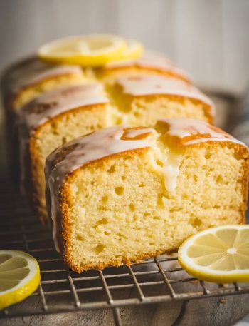Three thick slices of golden lemon pound cake arranged on a wire cooling rack with a glossy lemon syrup glaze on top and thin lemon rounds as garnish on a wooden surface