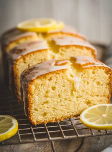 Three thick slices of golden lemon pound cake arranged on a wire cooling rack with a glossy lemon syrup glaze on top and thin lemon rounds as garnish on a wooden surface