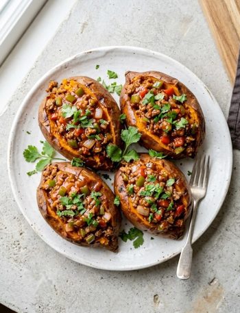 Four baked sweet potatoes sliced open and loaded with savory Whole30 paleo sloppy joe ground beef filling, garnished with fresh cilantro on a white plate