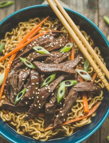 A deep blue ceramic bowl of slow cooker beef ramen noodles with tender glossy beef strips over golden ramen noodles topped with sliced green onions sesame seeds and julienned carrots with chopsticks resting on the bowl