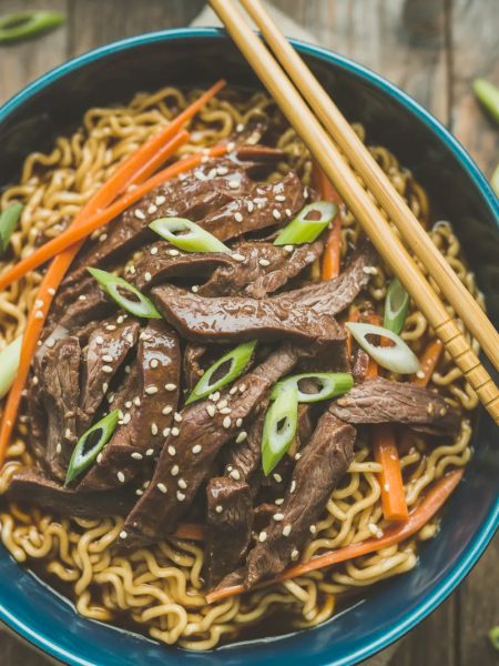 A deep blue ceramic bowl of slow cooker beef ramen noodles with tender glossy beef strips over golden ramen noodles topped with sliced green onions sesame seeds and julienned carrots with chopsticks resting on the bowl