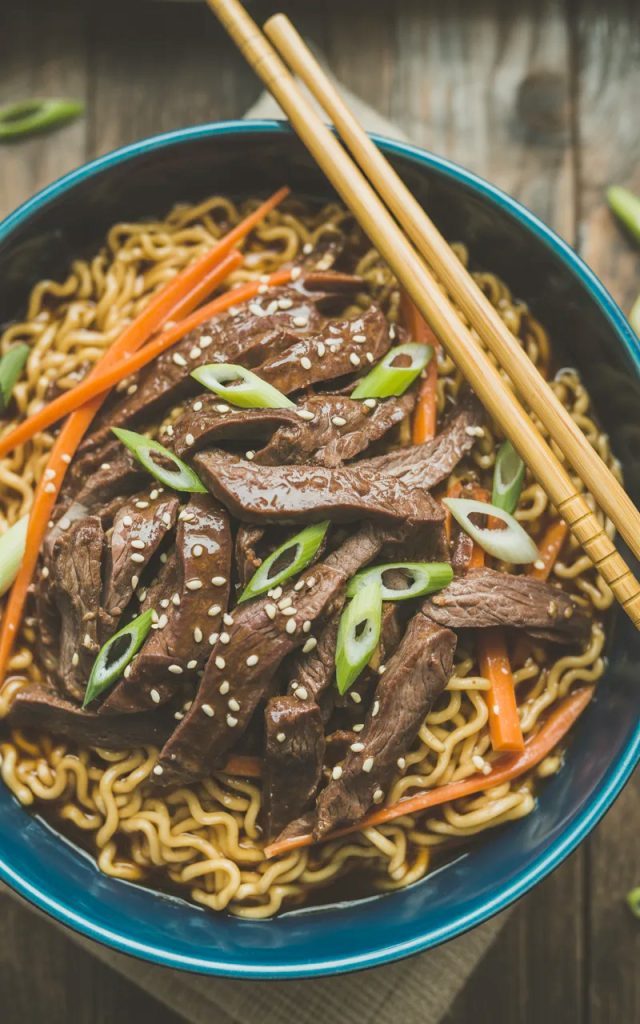 A deep blue ceramic bowl of slow cooker beef ramen noodles with tender glossy beef strips over golden ramen noodles topped with sliced green onions sesame seeds and julienned carrots with chopsticks resting on the bowl