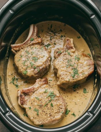 Three golden seared pork chops in a slow cooker smothered in a rich creamy garlic parmesan sauce garnished with fresh parsley and cracked black pepper