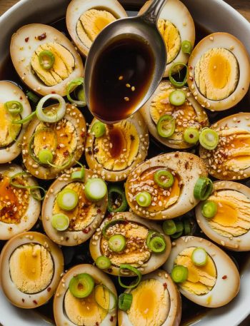 Overhead view of multiple halved soy marinated hard boiled eggs arranged in a white ceramic bowl showing deep amber stained egg whites and golden yolks, topped generously with sliced green onions, white sesame seeds, red chili flakes, and a spoon drizzling dark glossy marinade over the top