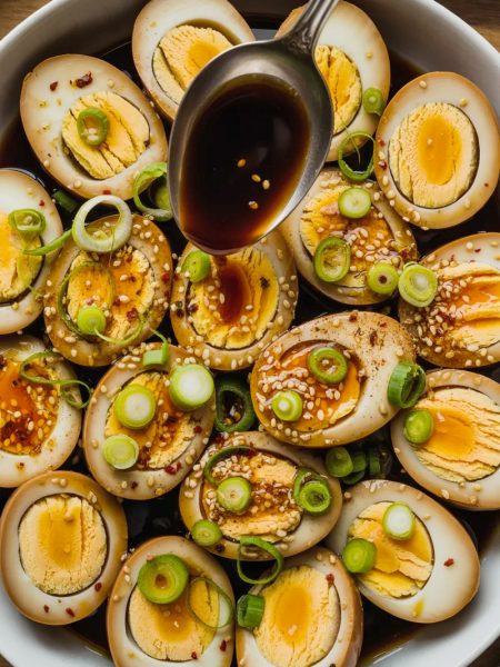 Overhead view of multiple halved soy marinated hard boiled eggs arranged in a white ceramic bowl showing deep amber stained egg whites and golden yolks, topped generously with sliced green onions, white sesame seeds, red chili flakes, and a spoon drizzling dark glossy marinade over the top