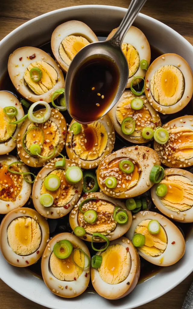 Overhead view of multiple halved soy marinated hard boiled eggs arranged in a white ceramic bowl showing deep amber stained egg whites and golden yolks, topped generously with sliced green onions, white sesame seeds, red chili flakes, and a spoon drizzling dark glossy marinade over the top