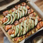 Spicy salmon sushi bake in a square baking dish topped with sliced avocado black sesame seeds green onions and zigzag drizzle of sriracha mayo and Kewpie mayo on a wooden surface