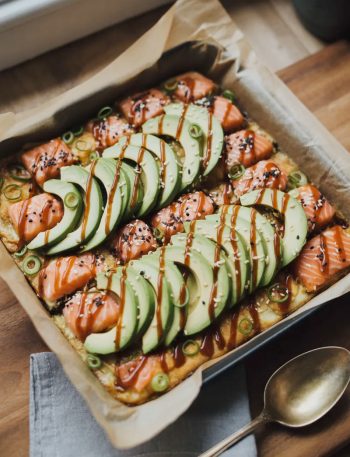 Spicy salmon sushi bake in a square baking dish topped with sliced avocado black sesame seeds green onions and zigzag drizzle of sriracha mayo and Kewpie mayo on a wooden surface