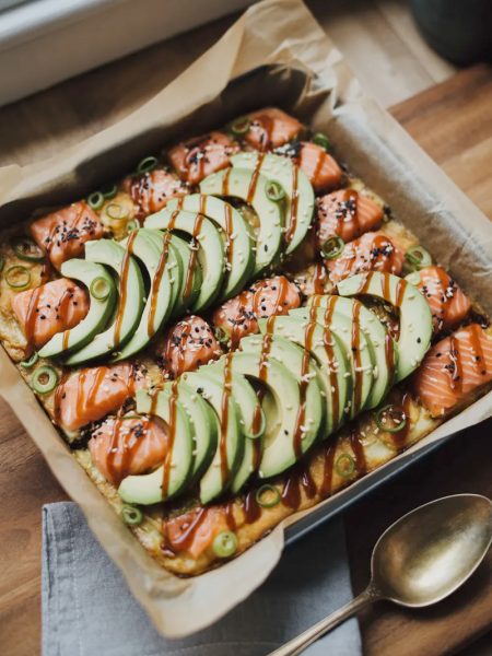Spicy salmon sushi bake in a square baking dish topped with sliced avocado black sesame seeds green onions and zigzag drizzle of sriracha mayo and Kewpie mayo on a wooden surface
