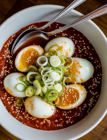 Overhead view of six soft boiled eggs sitting in a deep red spicy gochujang sauce in a white ceramic bowl, eggs lightly stained from the sauce, topped generously with sliced green onions and sesame seeds with two silver spoons resting in the bowl
